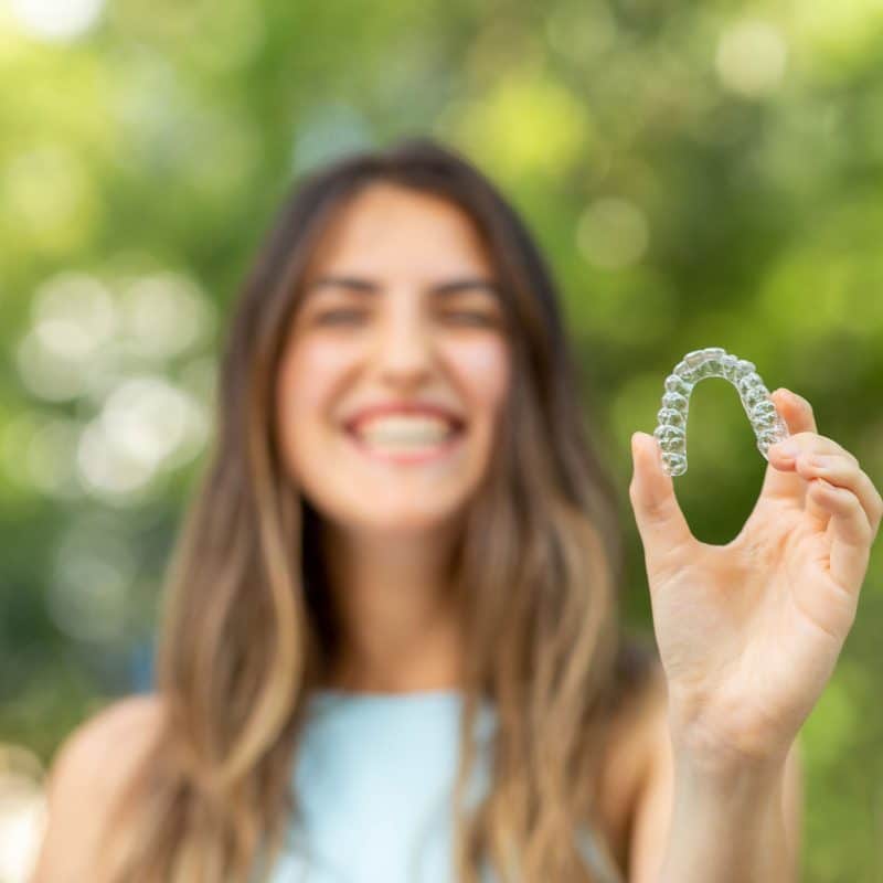 Woman holding clear dental aligner outdoors, smiling.