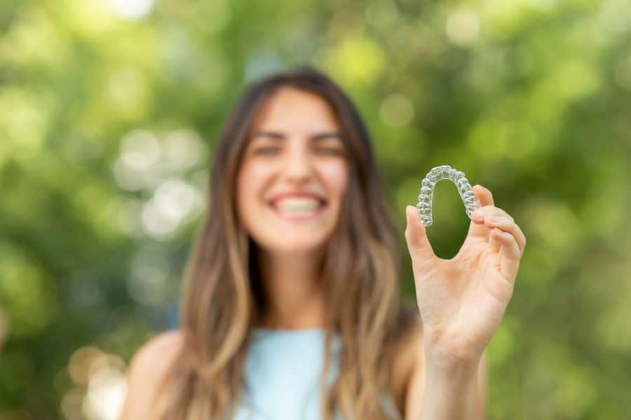 Woman holding clear dental aligner outdoors, smiling.